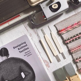 A flat lay showing a flat bed knitting machine, knitting machine tools and weights, a knitting swatch and a book called Designing Knitted Textiles
