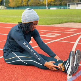Jimi sits on a running track with her legs stretched out straight in front of her and her arms reaching towards her ankles