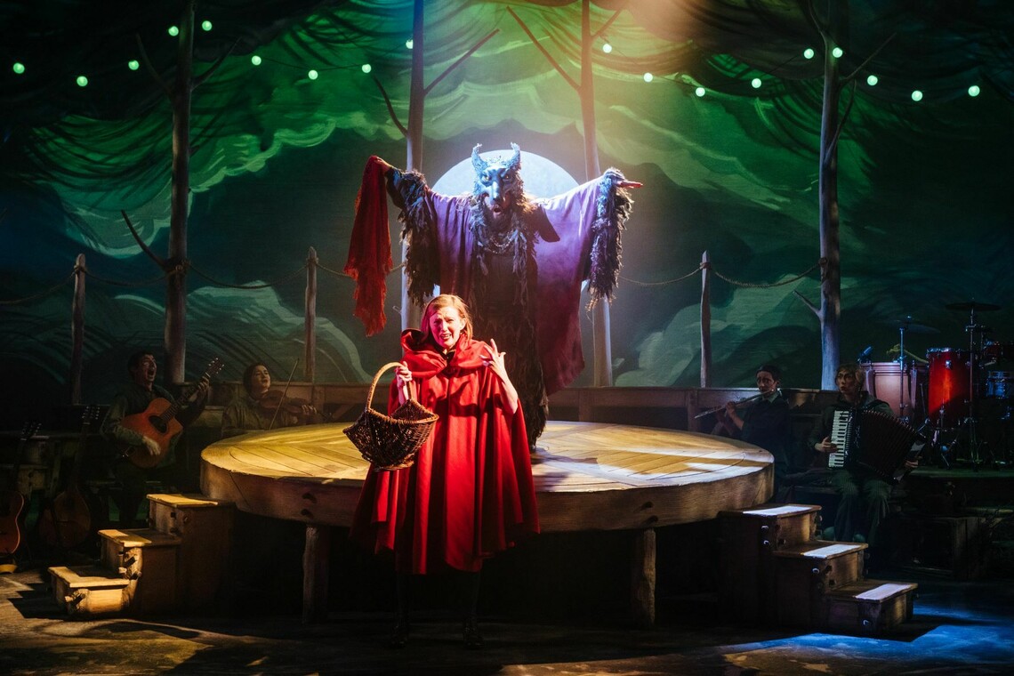 A woman dressed in a red cloak with a puppet wolf behind