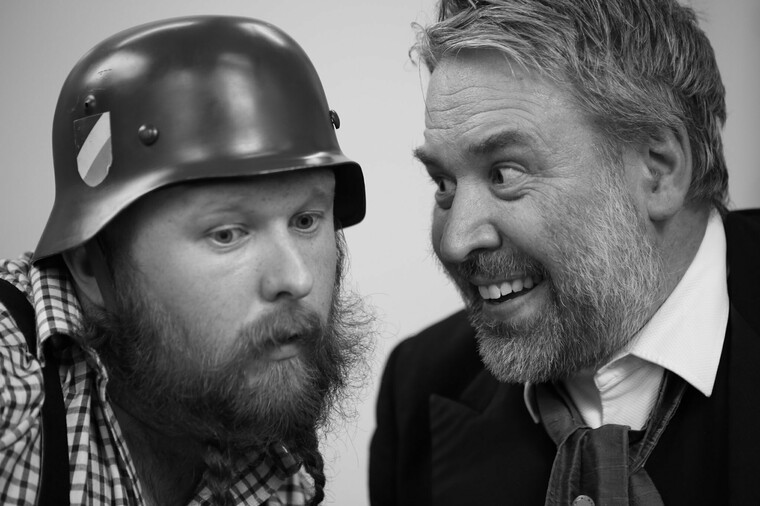 Two male actors, one grinning, the other wearing a helmet leaning in