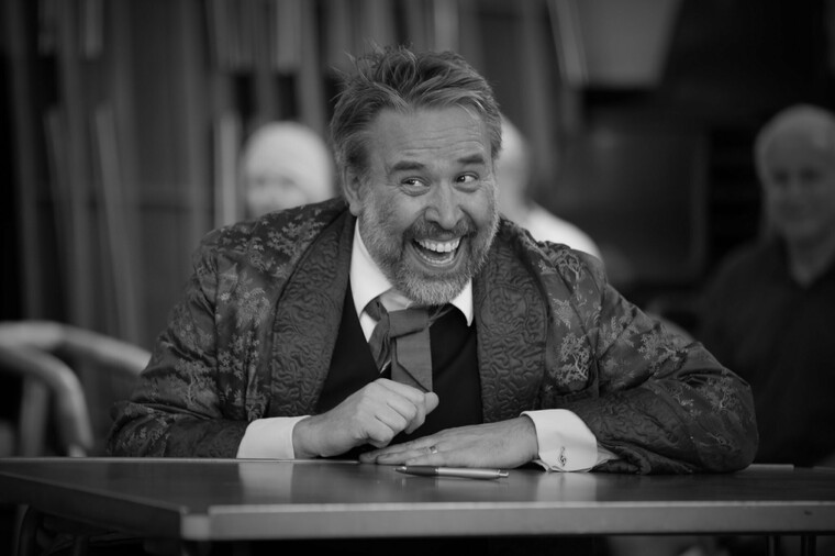 Male actor, seated with arms on table and a broad grin