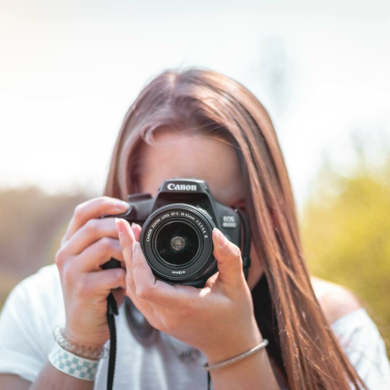 Girl with long brown hair and camera held obscuring her face