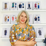 Blond middle aged lady in brightly coloured geometric patterned shirt standing in front of various patterns displayed in the background on shelves
