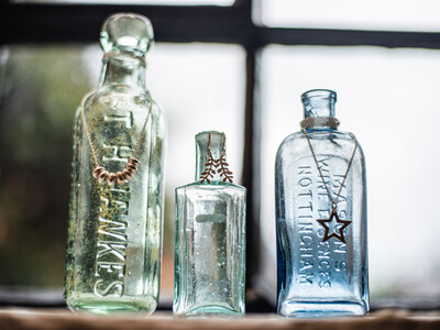 three different sized vintage glass bottle standing on a window sill each with jewellery hanging form the bottle necks