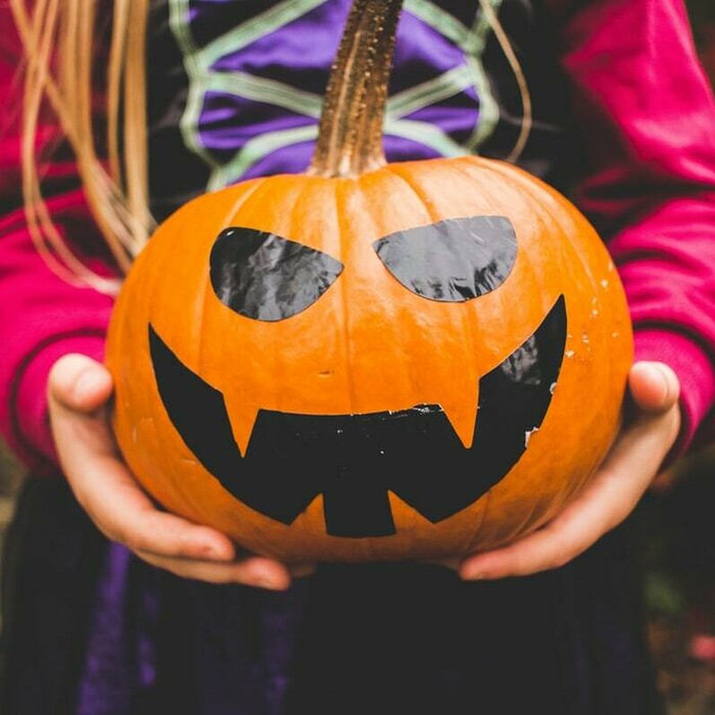 A young girl in a Halloween costume holds a decorated Halloween pumpkin.