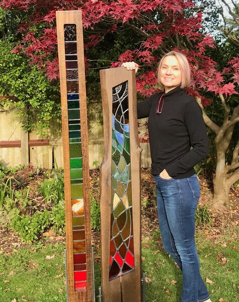 A blonde woman stands in a garden with her arm resting on one of two glass mosaic sculptures.