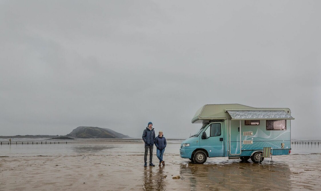 A camper van parked on the sand of a beach with a couple stood next to it