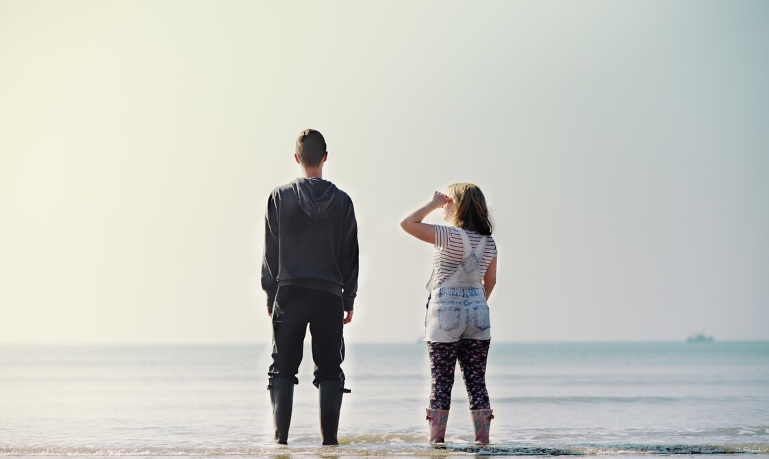 Two young people stand on a beach with their backs to the camera, looking out to sea.