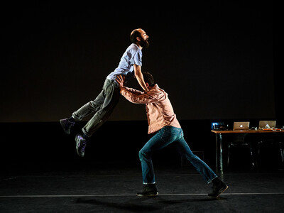 One man in jeans and a pink shirt lifts another man in to the air.