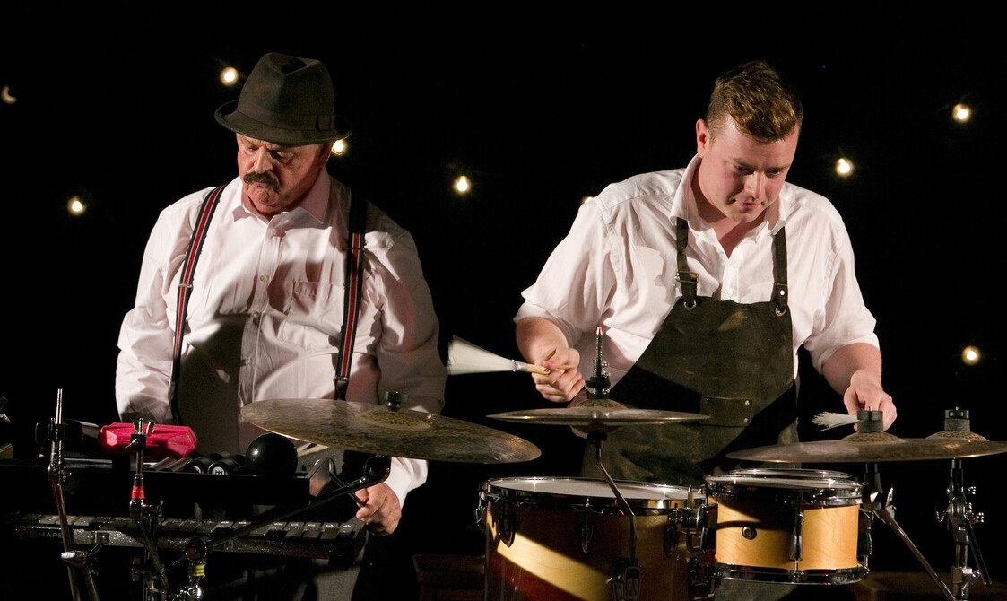 Two actors standing side by side playing the drums and the xylophone.