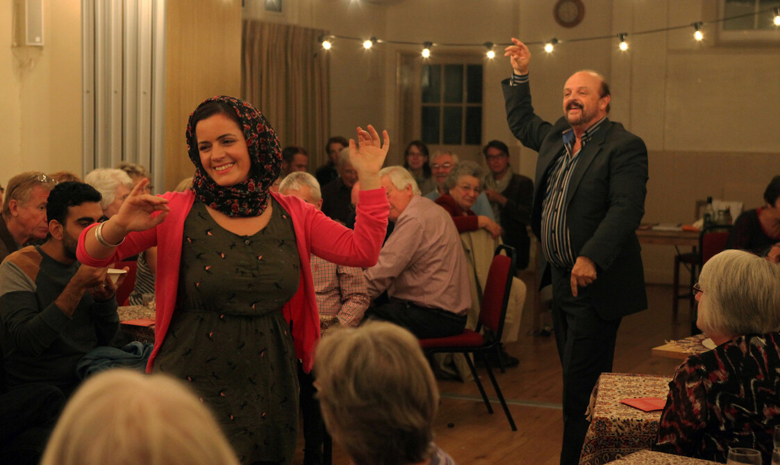 Two actors dance in the middle of a village hall, surrounded by audience members sitting at tables.