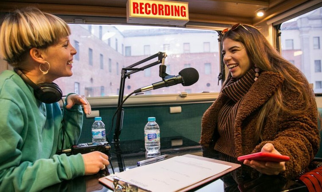 Two woman in a recording studio talk into a microphone.