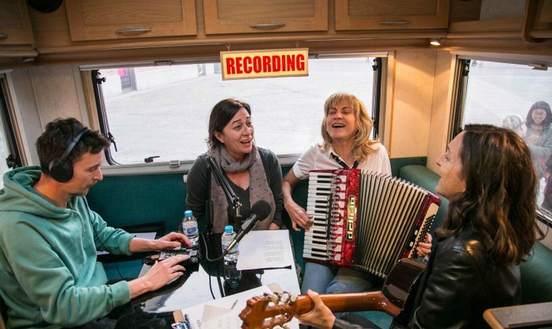 A group of people with instruments sit in a recording studio talking and playing their instruments.