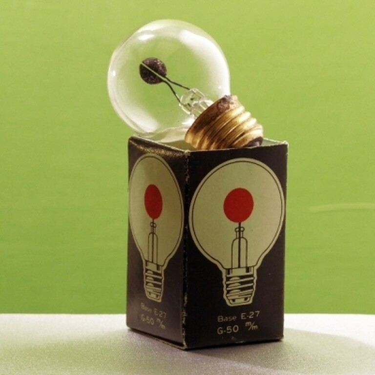 A filament light bulb resting on a light bulb box in front of a green wall