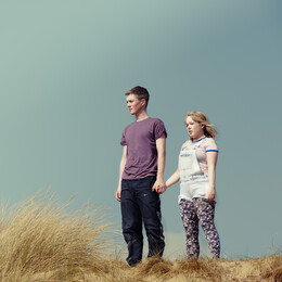 A young man and woman stand holding hands on a beach sand dune