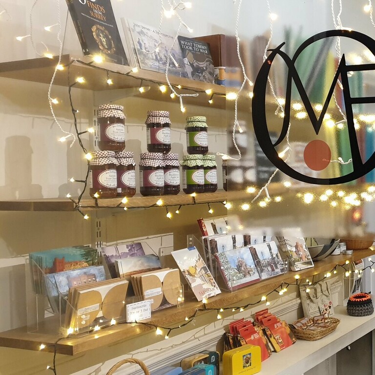 A view through the window of the museum of farnham shop with products displayed on shelves and fairy lights