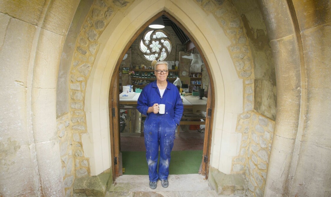 A person stands in a church archway in blue over-all's holding a cup of tea