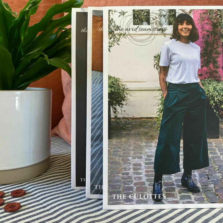 3 sewing patterns packets stood up right with the front one showing a photos of a lady on a mediteranian patio with green culottes on. a plant pot in the background and terracotta walls.
