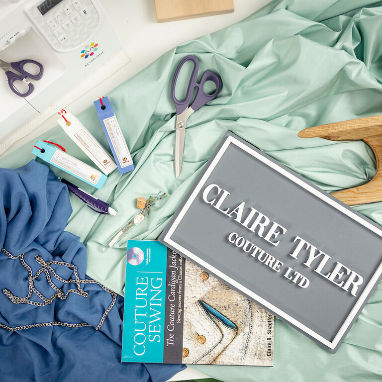 A table covered in fabric, scissors, a sewing machine, a chain and a sign that reads: Claire Tyler