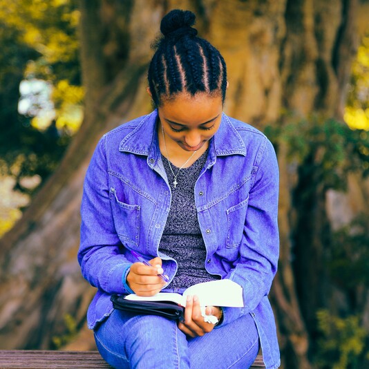 A young woman sits outside by a tree writing into a journal