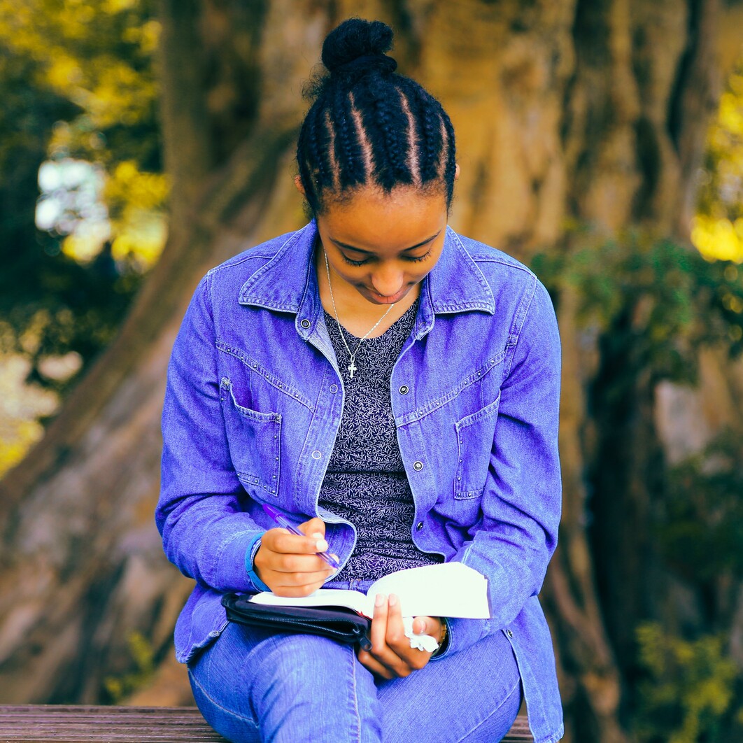 A young woman sits outside by a tree writing into a journal
