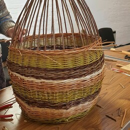 Partially woven basket on table with hand threading next twine though.  Top part of basket still to be finished