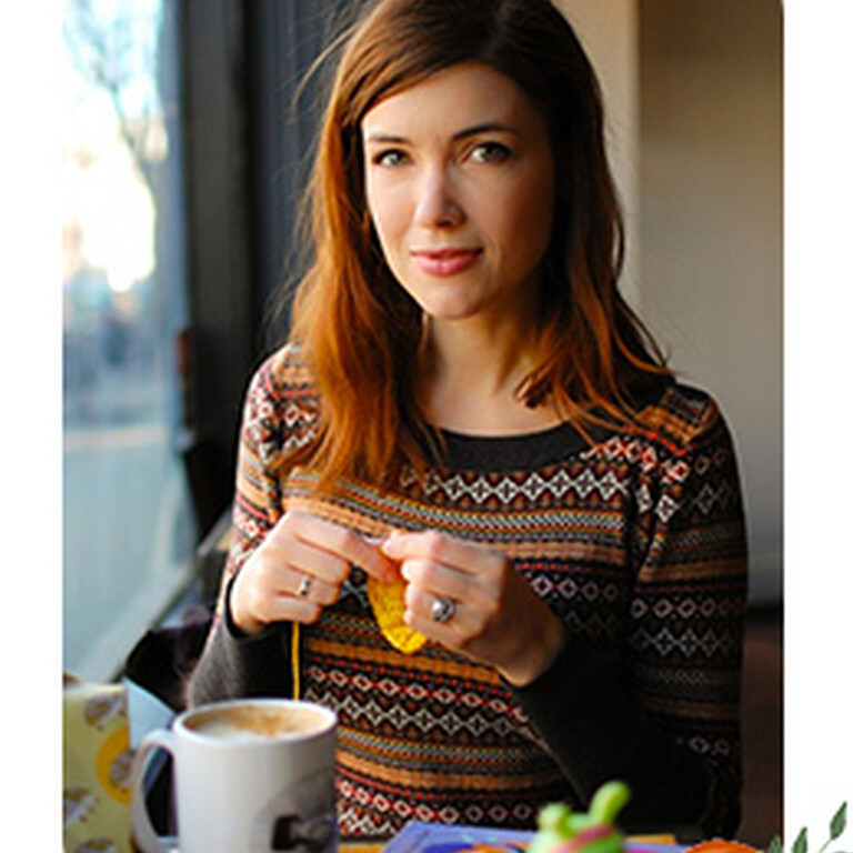 Photo of Julie Crawford knitting sat at a table with a cup of tea.