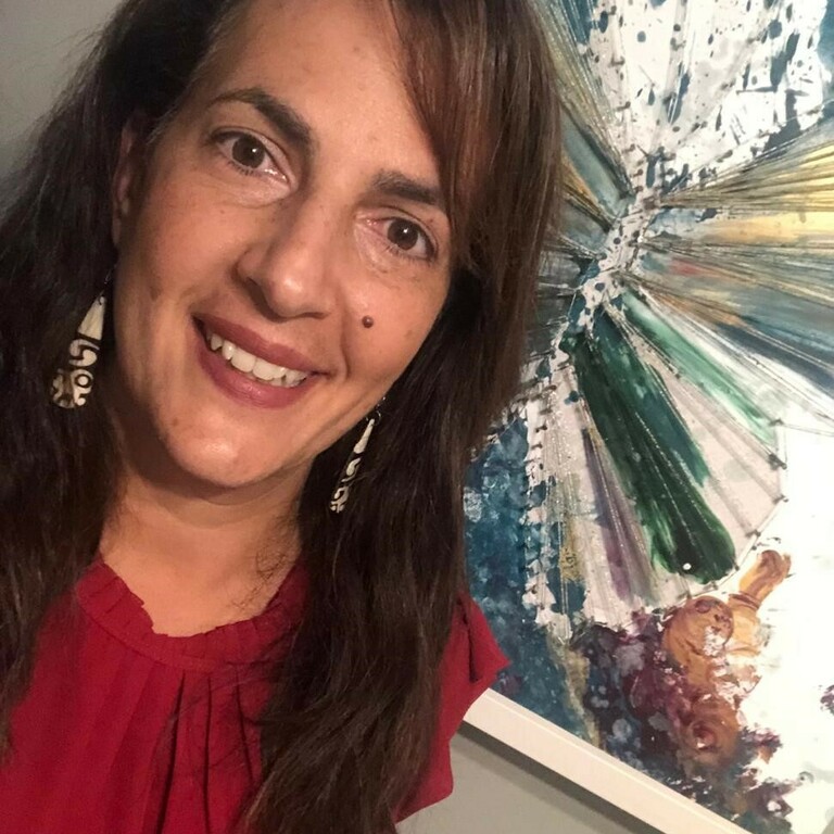 A head shot of craft tutor Nicole Marcussen standing in front of a painting that she painted, hung on a wall