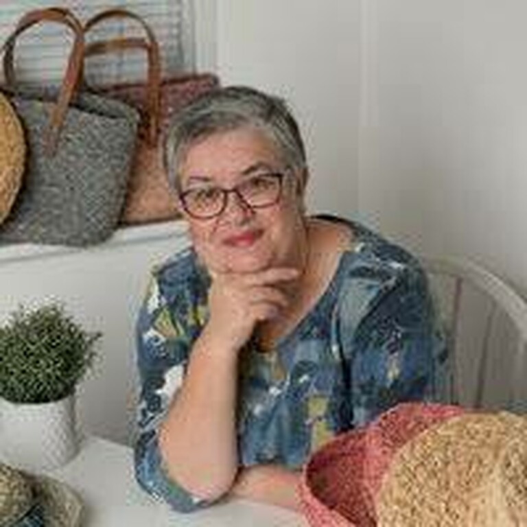 A woman with glasses wearing a blue print top. Behind her are raffia bags and in front of her are raffia hats.
