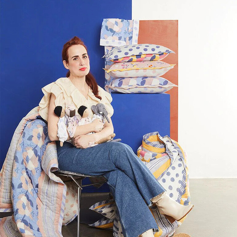 A woman sits holding four fabric dolls. Around her are quilted blankets and cushions