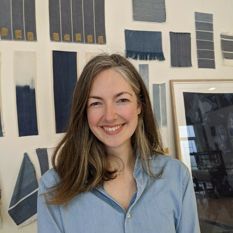 a lady in a blue shirt smiling stood in front of some fabric art work in the back ground