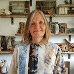 A lady with a hand printed shirt on in mottled blues and rust colours stood in front of shelves with interesting craft, ceramics and vintage notions.