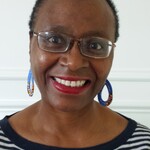 A black woman wearing glasses, a stripy top and multicoloured earrings, smiling.