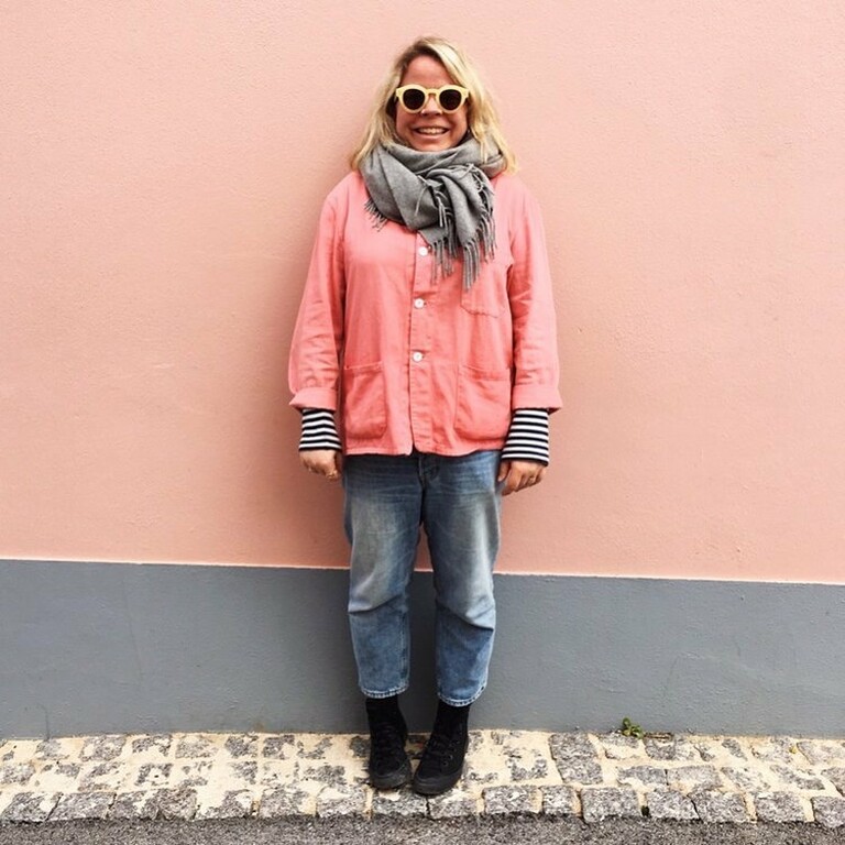 Lucy Rowan Wearing a pink jacket, with a scarf and sunglasses leaning on a pink wall