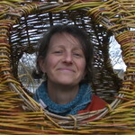A photo o Judith Needham peering her head out of a woven willow structure.