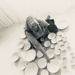 A black and white photo of Penny Eccles sat on the floor surrounded by ceramic dishes