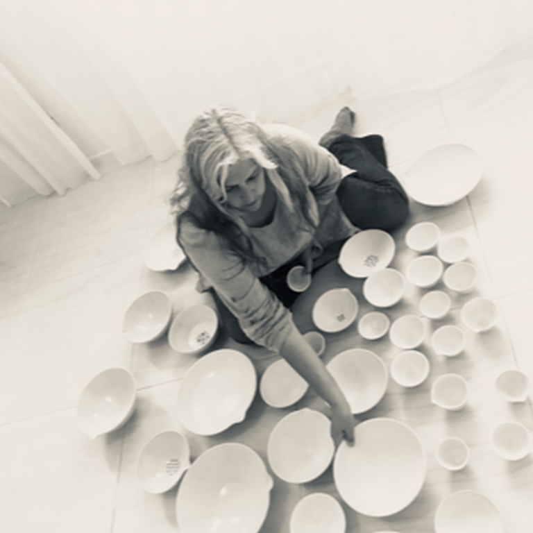 A black and white photo of Penny Eccles sat on the floor surrounded by ceramic dishes