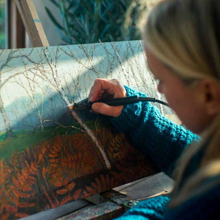 Artist Ruth Wheeler holding a heated metal pen, burns details into a woodland painting.