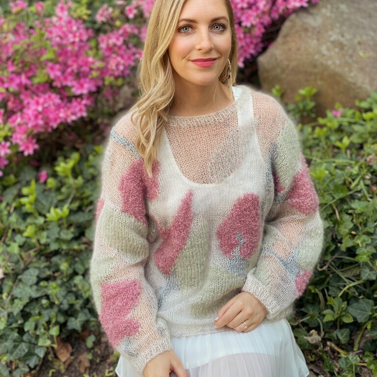 A smiling blonde woman sits on a rock outside wearing a white dress and white and pink jumper.