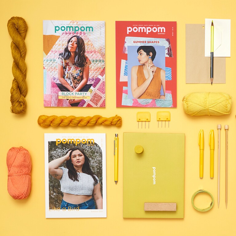a flat lay on a yellow background with three issues of Pom Pom Quarterly surrounded by writing instruments, skeins of wool and knitting needles