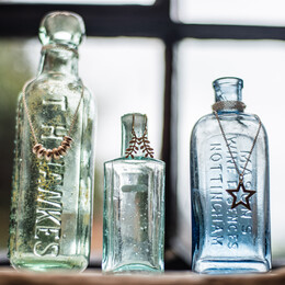 three different sized vintage glass bottle standing on a window sill each with jewellery hanging form the bottle necks