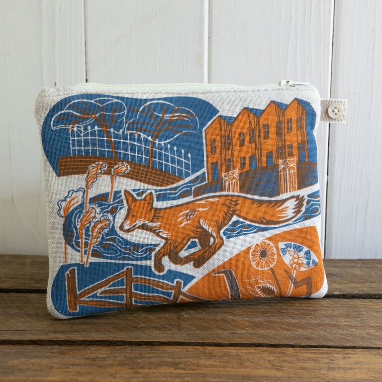 A white cushion with a design of a fox running through town, on a wooden table.
