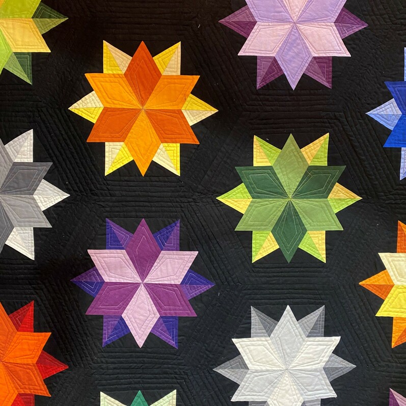A black logo with multicoloured star motifs for Quilters Fair 2022