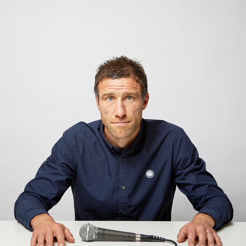 Image of Andrew at a desk wearing a blue jumper, with a microphone placed in front of him