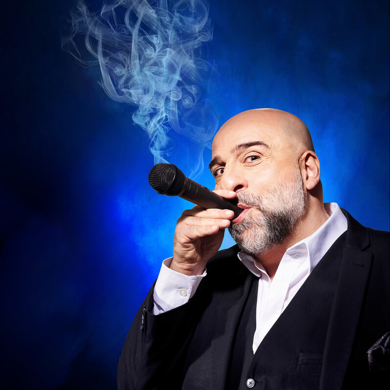 Promotion shot of comedian Omid Djalili imitating smoking a microphone