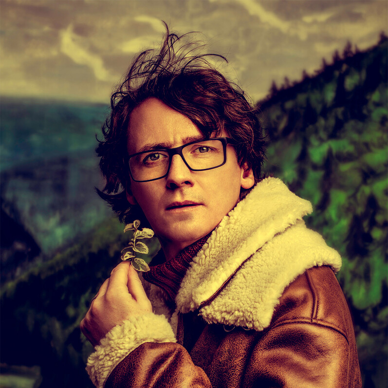 Promotion shot of comedian Ed Byrne standing against a backdrop of a mountain, holding a plant