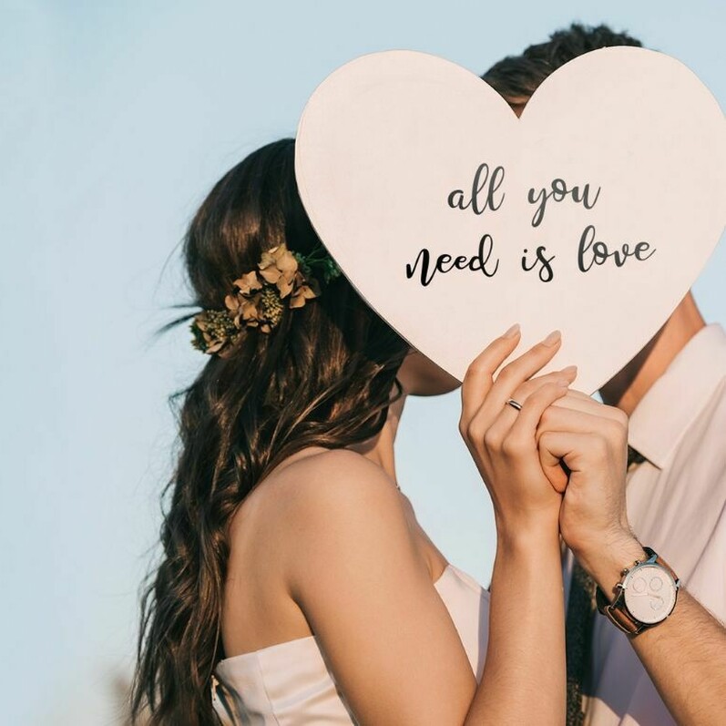 Man and woman hide their faces behind love heart-shaped sign saying all you need is love