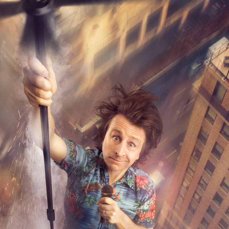 Milton Jones promotional image showing him in the air holding a propeller and a microphone above buildings