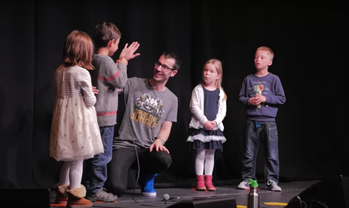 A man stands on stage with a group of children and hand fives a young boy.