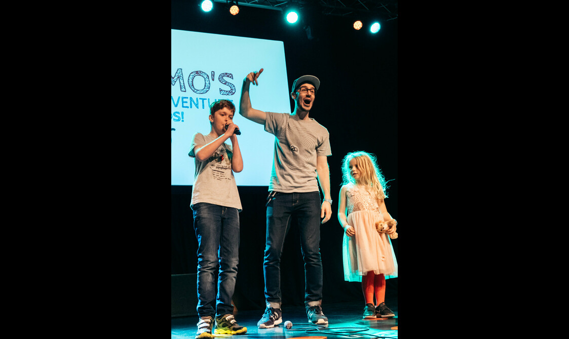 A man hypes up the audience standing between two children on stage as they Beat box